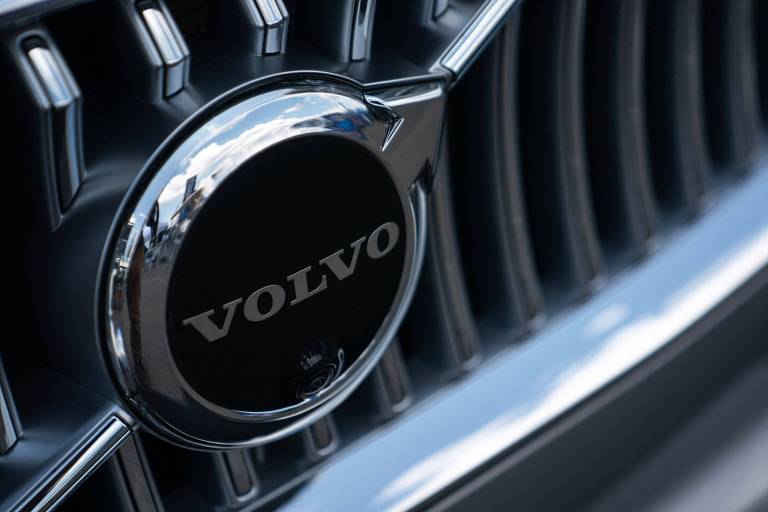 A imagem mostra um close do logotipo da Volvo, que é circular e apresenta a palavra 'VOLVO' em letras brancas sobre um fundo preto. O logotipo está inserido em uma grade de carro, com detalhes metálicos ao redor.
