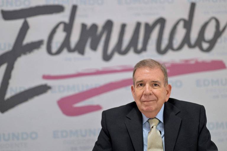 Um homem de cabelos grisalhos e pele clara está sentado em uma mesa, vestindo um terno escuro e uma gravata clara. Ao fundo, há uma parede com o nome 'Edmundo' escrito em letras grandes e estilizadas, em cores que variam entre preto e rosa.