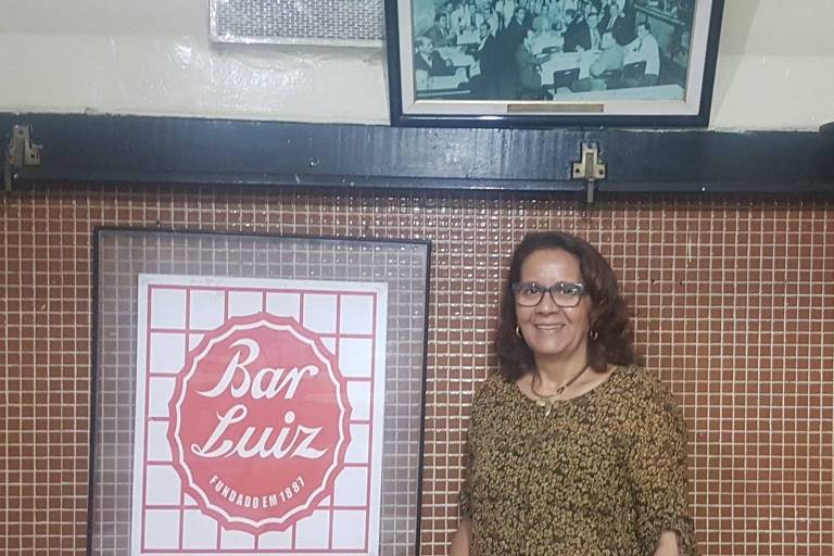 Uma mulher está em pé ao lado de um quadro com o logotipo do Bar Luiz, que é um círculo vermelho com o nome 'Bar Luiz' escrito em branco. Ao fundo, há uma foto em preto e branco emoldurada, mostrando um grupo de pessoas em um ambiente que parece ser um bar ou restaurante. A mulher usa óculos e uma blusa de estampa animal.