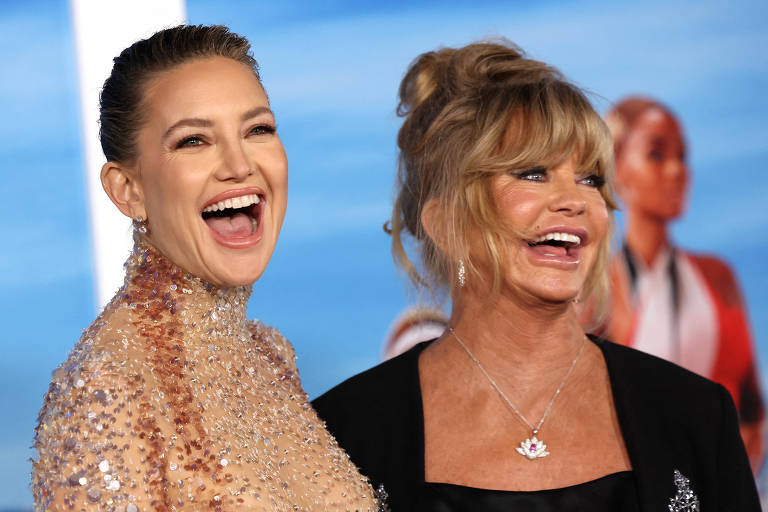 Duas mulheres estão sorrindo amplamente em um evento. A mulher à esquerda usa um vestido brilhante e tem o cabelo preso, enquanto a mulher à direita está com um vestido preto e cabelo solto, com um coque bagunçado. Ambas parecem felizes e estão em um ambiente com fundo azul.