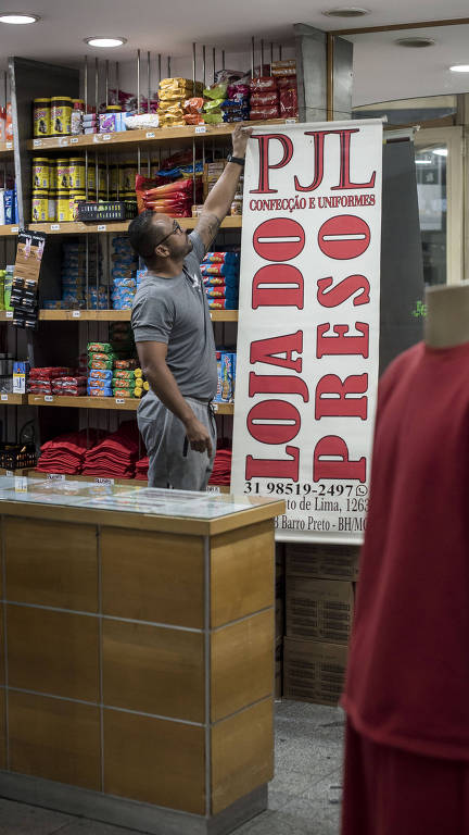 A imagem mostra um homem em uma loja, organizando produtos em uma prateleira. Ao fundo, há uma placa grande com o nome 'LOJA DO PRESO' em letras vermelhas. O ambiente é bem iluminado e as prateleiras estão cheias de mercadorias, incluindo roupas e outros itens. Um manequim com uma camiseta vermelha está em primeiro plano.