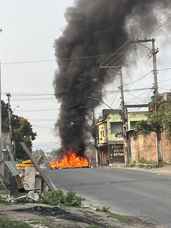 Imagem colorida mostra uma rua do Complexo do Salgueiro, em São Gonçalo, na Região Metropolitana do Rio, bloqueada por um carros incendiado; é possível ver muita fumaça preta saindo do veículo, e mais à frente um monte de entulho também usado como barricada