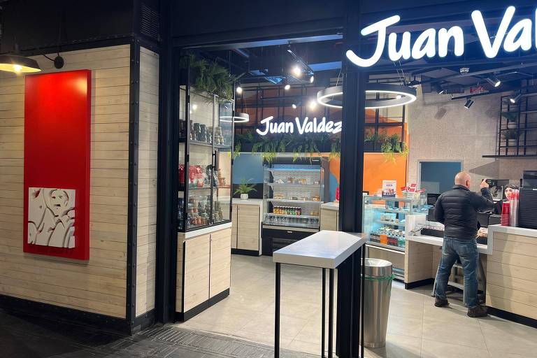 A imagem mostra a entrada de uma cafeteria Juan Valdez. O espaço tem paredes de madeira clara e um painel vermelho com uma escultura em relevo. Dentro, há prateleiras com produtos expostos e um balcão onde um cliente está sendo atendido. O ambiente é iluminado com luzes pendentes e há plantas decorativas visíveis.