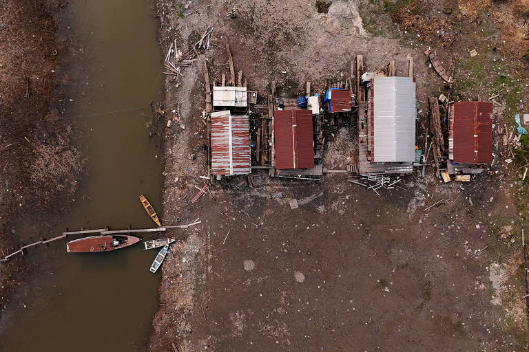 A imagem mostra uma vista aérea de várias estruturas de madeira com telhados vermelhos, localizadas próximas a um corpo d'água. À esquerda, há um barco ancorado e uma pequena ponte de madeira que conecta as estruturas ao solo. O terreno ao redor é seco e apresenta detritos, com vegetação escassa visível nas bordas.