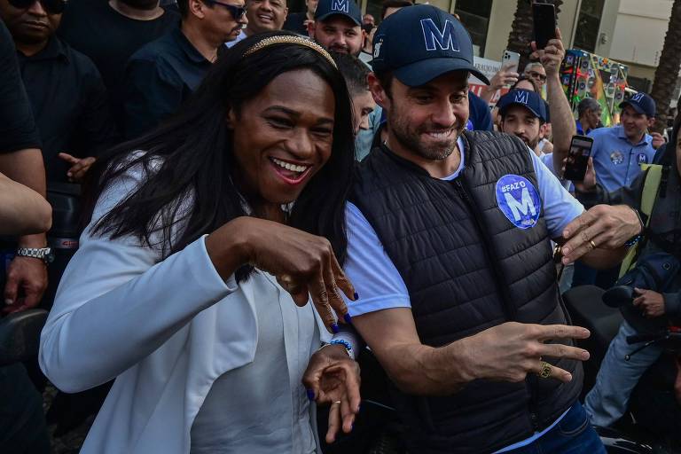 Pablo Marçal é um homem branco de barba baixa e escura. Ele veste um boné azul escuro com a letra M bordada, uma camiseta azul clara e uma jaqueta puffer sem mangas com um adesivo com a letra M colado. Ele morde os lábios e estende três dedos em cada mão, simulando a letra M. Antônia de Jesus é uma mulher negra com cabelos escuros abaixo dos ombros. Ela veste um casaco de cor clara e uma tiara. Está à esquerda de Marçal e faz os mesmos gestos que ele. Eles estão rodeados de várias pessoas