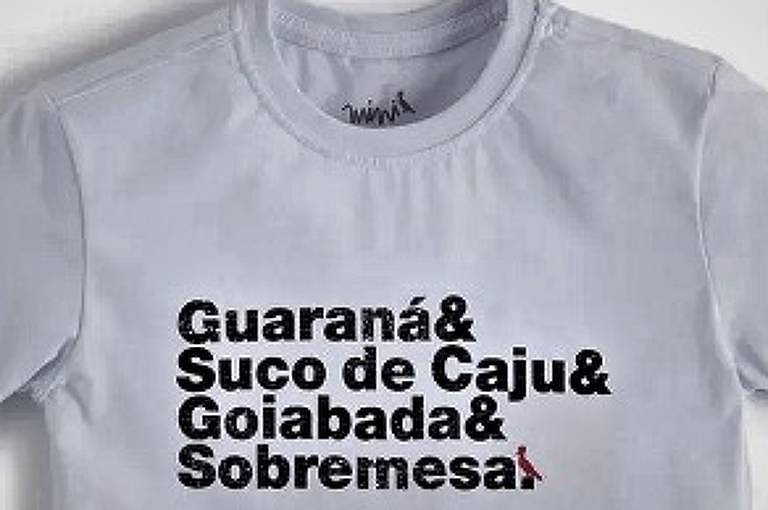 A imagem mostra uma camiseta de cor clara com um design gráfico em preto. O texto na camiseta diz: 'Guaraná& Suco de Caju& Goiabada& Sobremesa&'.