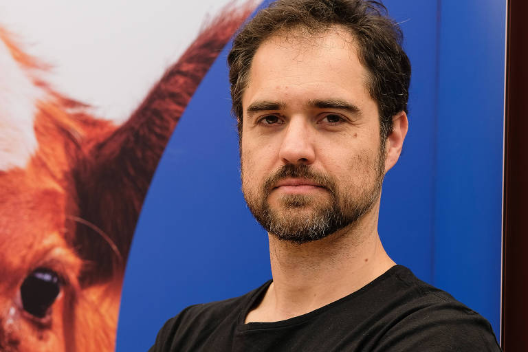 Um homem está posando com os braços cruzados. Ele tem cabelo escuro e barba. Está usando uma camiseta preta com a palavra 'Liberade' e um desenho colorido. Ao fundo, há uma imagem de uma vaca e um fundo azul.