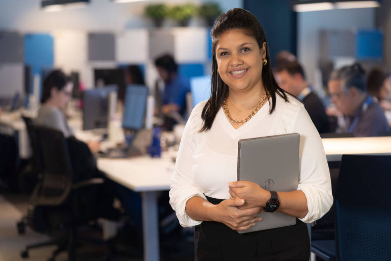 A imagem mostra uma mulher sorridente em um ambiente de escritório moderno. Ela está segurando um laptop e usa uma blusa branca com mangas bufantes e uma calça preta. Ao fundo, há várias pessoas trabalhando em mesas com computadores. O ambiente é bem iluminado e decorado com plantas.