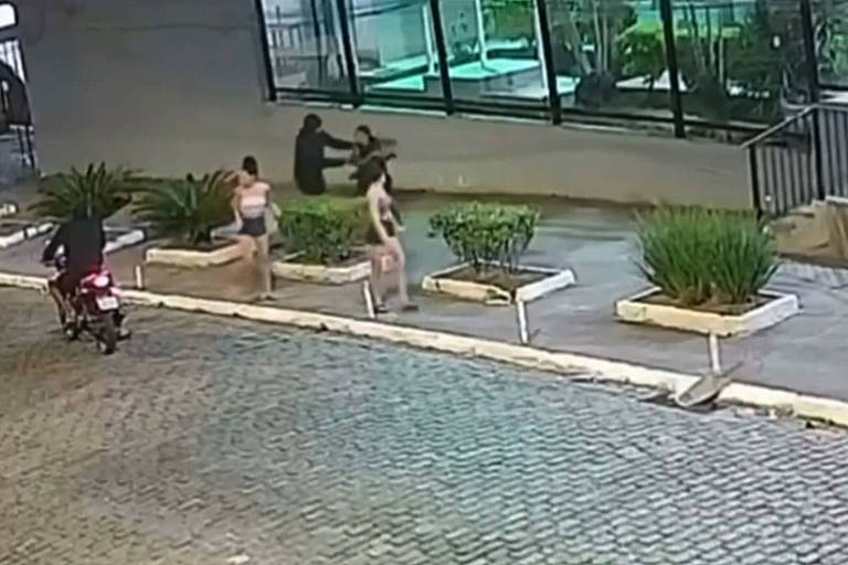 A imagem mostra uma área externa com um pavimento de pedras. À esquerda, um motociclista está parado. No centro, duas mulheres estão em pé, enquanto um homem segura nos braços da jovem perto de uma parede. Ao fundo, há uma estrutura de vidro e algumas plantas em vasos.