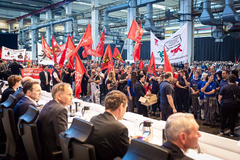 A imagem mostra uma grande manifestação de operários da Volkswagen, com várias pessoas segurando bandeiras vermelhas e cartazes. No primeiro plano, está a diretoria da empresa com pesoas sentadas em uma mesa, aparentemente em uma posição de destaque, observando a manifestação. O ambiente é industrial, com estruturas metálicas visíveis ao fundo.