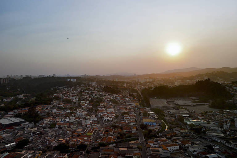 A imagem mostra uma vista panorâmica de uma cidade ao entardecer, com o sol se pondo no horizonte. A cidade é composta por uma densa aglomeração de casas e edifícios, com telhados de diferentes cores, principalmente tons de laranja e cinza. Ao fundo, há colinas e uma leve neblina, criando um efeito suave na paisagem. O céu apresenta um gradiente de cores que vai do azul ao laranja, indicando o final do dia.