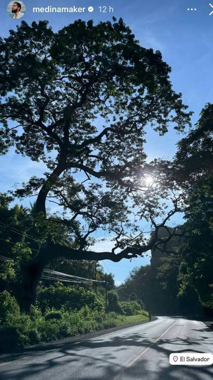 A imagem mostra uma estrada em um ambiente natural, com uma grande árvore à esquerda, cujos galhos se estendem sobre a via. O céu está claro, com algumas nuvens, e o sol brilha ao fundo, criando um efeito de luz. À direita, há um ciclista pedalando na estrada. O local é identificado como El Salvador.