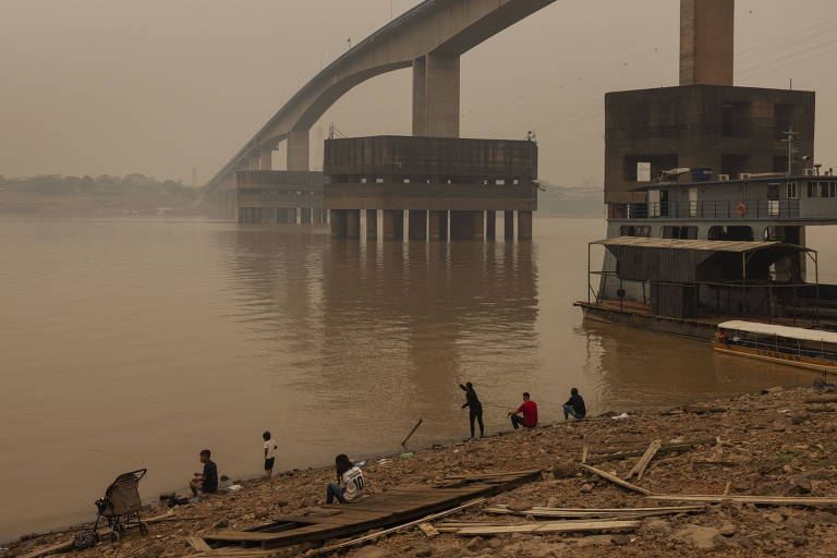 A imagem mostra uma ponte elevada sobre um rio, com uma estrutura de concreto ao lado. A água do rio Madeira está turva e há uma névoa densa no ar, indicando poluição. Algumas pessoas estão na margem do rio, aparentemente pescando ou coletando algo. O cenário é sombrio, com cores apagadas devido à poluição.