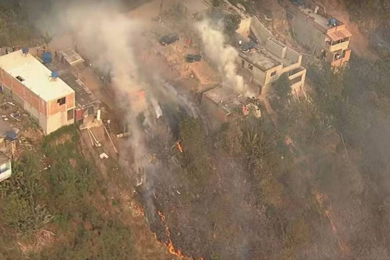 A imagem mostra uma área urbana afetada por um incêndio. Há fumaça saindo de várias partes, e chamas visíveis em uma área de vegetação. Algumas casas estão visíveis, com telhados de diferentes cores, e há veículos estacionados nas proximidades. A vegetação ao redor parece queimada e danificada pelo fogo