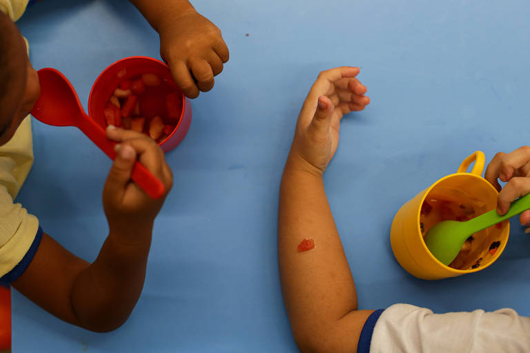 A imagem mostra as mãos de duas crianças segurando colheres e comendo de tigelas coloridas. Uma criança está usando uma colher vermelha e a outra uma colher verde. As tigelas são de cores rosa e amarela, e a mesa é coberta por um pano azul