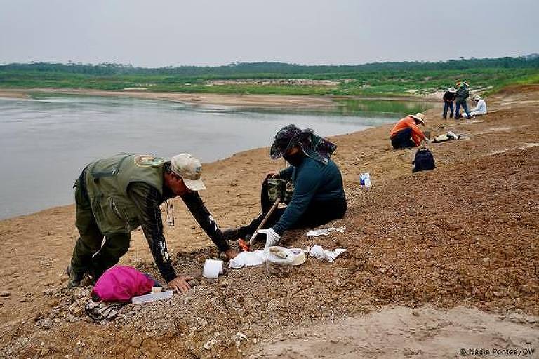 A imagem mostra várias pessoas trabalhando na beira de um rio. Algumas estão agachadas, mexendo na areia ou em objetos, enquanto outras estão sentadas. O ambiente é natural, com vegetação ao fundo e um céu nublado. O rio está calmo e a areia é clara.