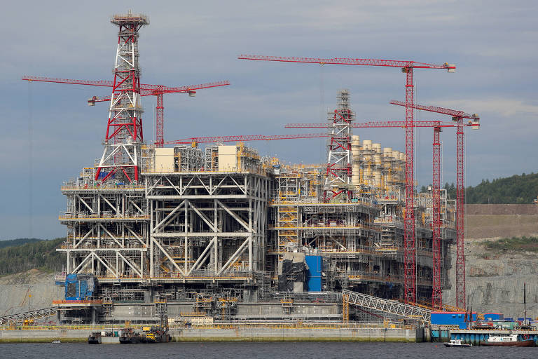 A imagem mostra uma grande estrutura de construção industrial, possivelmente uma plataforma de petróleo ou gás, com várias gruas vermelhas ao redor. A estrutura é composta por uma base metálica complexa e está localizada à beira de um corpo d'água. O céu está parcialmente nublado, e ao fundo, é possível ver uma área de terreno com vegetação.