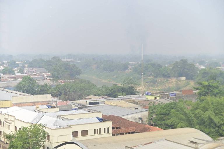 A imagem mostra uma vista de uma área urbana com prédios e vegetação. No fundo, há uma fumaça visível no céu, indicando poluição. O rio pode ser visto à direita, cercado por árvores. A atmosfera parece nebulosa devido à fumaça