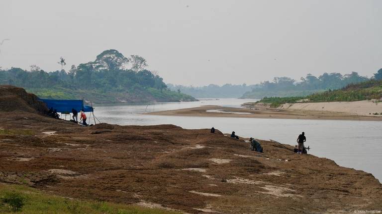 A imagem mostra uma cena à beira de um rio, com um grupo de pessoas trabalhando na margem. À esquerda, há uma estrutura coberta por uma lona azul. O rio se estende ao fundo, cercado por vegetação e uma atmosfera nevoenta. A margem do rio é composta por areia e pedras, e algumas pessoas estão visíveis, possivelmente pescando ou coletando algo.