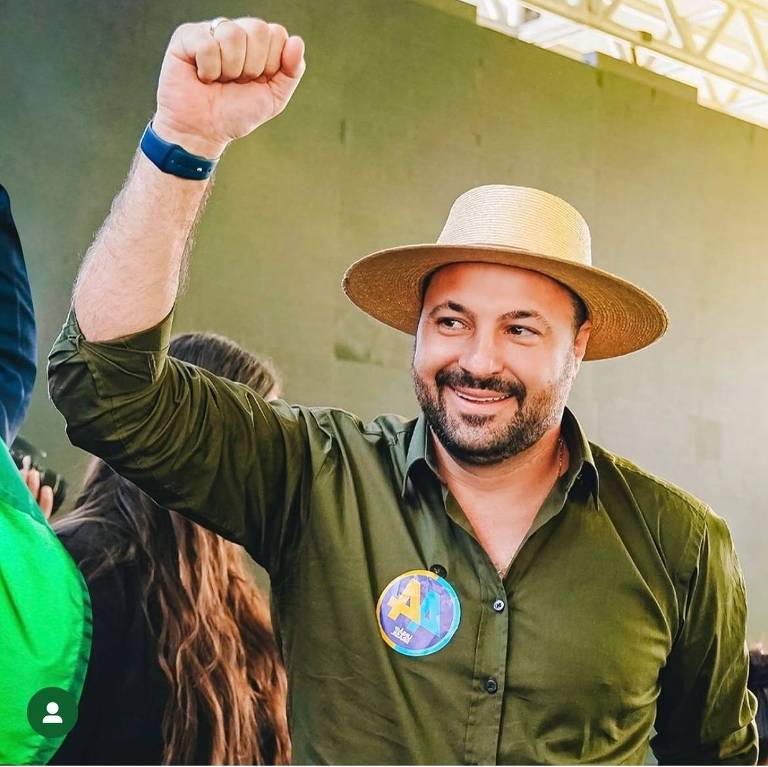 Um homem sorridente levanta o braço direito com o punho cerrado, usando uma camisa verde e um chapéu de palha. Ele está em um evento, cercado por outras pessoas, e parece estar celebrando ou se engajando em uma atividade animada.