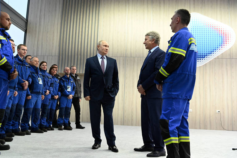 A imagem mostra um grupo de homens em uniformes azuis, possivelmente trabalhadores de uma empresa, alinhados em uma sala moderna. À frente deles, dois homens em trajes formais estão conversando. Um deles parece ser um líder ou figura importante, enquanto o outro escuta atentamente. O ambiente é bem iluminado, com paredes de madeira e um painel decorativo ao fundo.