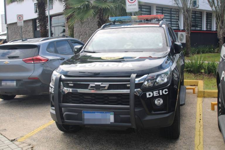 A imagem mostra uma viatura da polícia estacionada em uma área urbana. O veículo é preto com detalhes em branco e possui a palavra 'POLÍCIA' escrita na parte superior do capô. A viatura tem um emblema e a sigla 'DEIC' na lateral. No teto, há luzes de emergência vermelhas e azuis