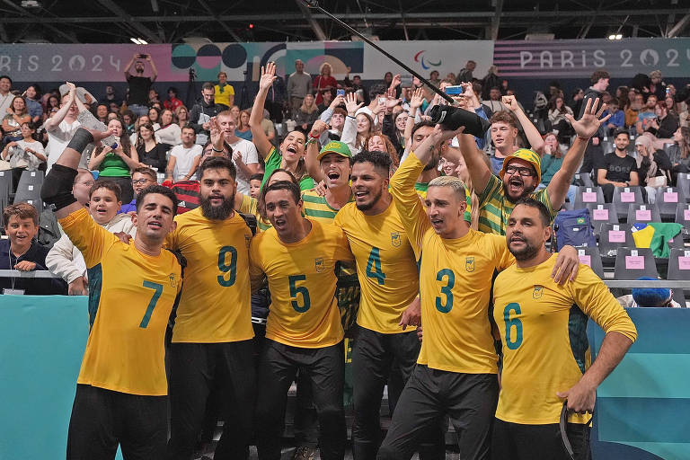 Um grupo de oito homens, todos vestindo camisetas amarelas com detalhes verdes, está posando para a foto em um ambiente de celebração. Eles estão sorrindo e levantando os braços, demonstrando alegria. Ao fundo, há uma multidão de torcedores, alguns deles também estão animados e aplaudindo. O ambiente parece ser uma arena esportiva, possivelmente durante um evento relacionado aos Jogos Paralímpicos de Paris 2024.
