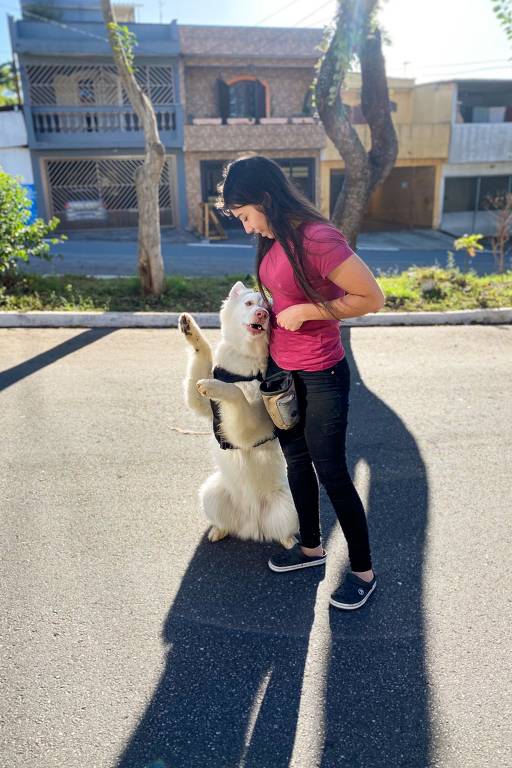 Uma mulher está em pé em uma rua, olhando para um cachorro que está em pé sobre as patas traseiras. O cachorro é de pelagem branca e parece estar feliz. A mulher usa uma camiseta rosa e calças escuras, e está segurando um objeto na cintura. Ao fundo, há casas e árvores, e a luz do sol ilumina a cena.