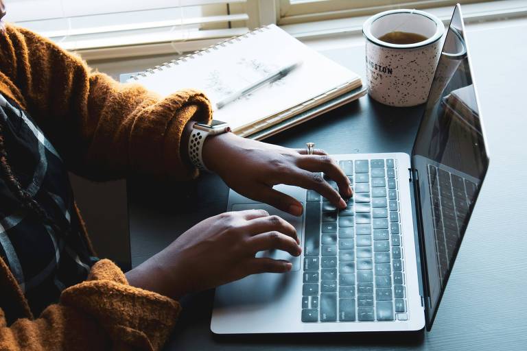 Mulher digitando em laptop. Ao lado, há um caderno aberto, uma caneta e uma xícara de café