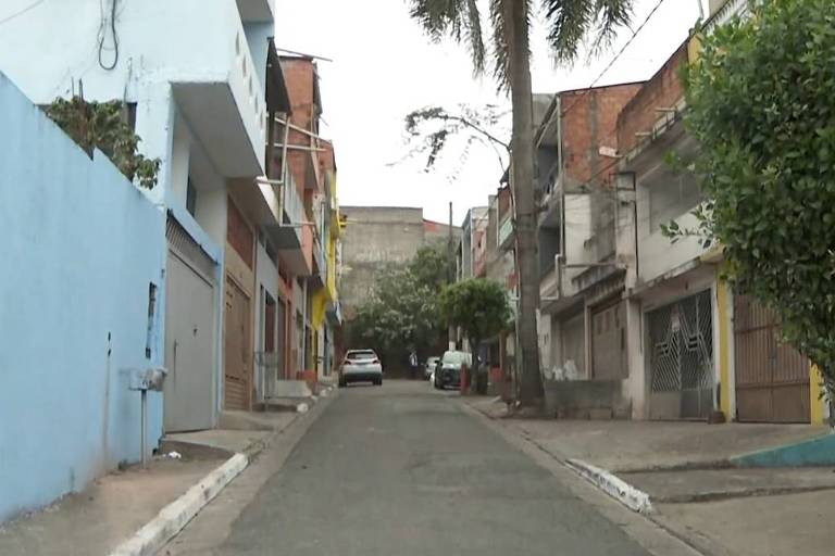 A imagem mostra uma rua em uma área urbana, com casas de diferentes cores e estilos ao longo dos dois lados. À esquerda, há uma parede azul e uma garagem. À direita, as casas são de alvenaria, algumas com grades nas janelas