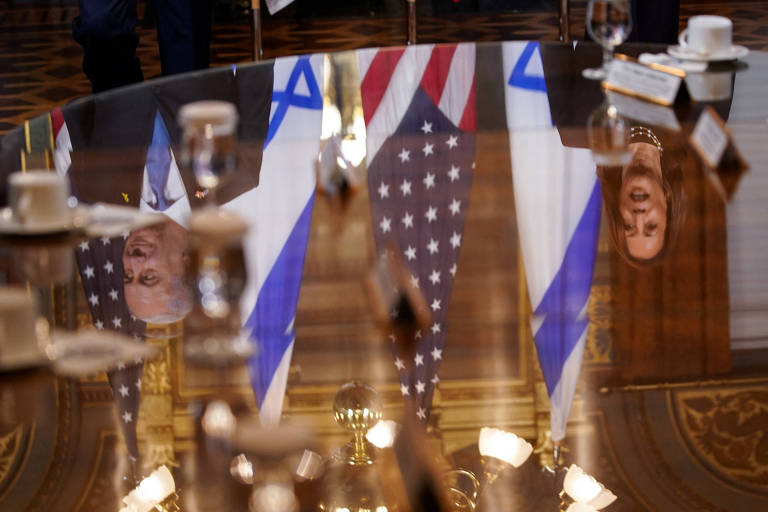 A imagem mostra uma mesa de reunião com um reflexo de bandeiras de Israel e dos Estados Unidos. As bandeiras estão dispostas verticalmente, com a bandeira dos Estados Unidos à direita e a de Israel à esquerda. O ambiente é formal, com copos e xícaras sobre a mesa. O reflexo das bandeiras é visível na superfície da mesa.