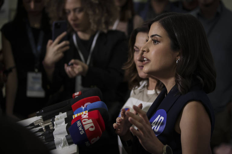 Fotografia mostra a deputada federal e candidata à Prefeitura de São Paulo Tabata Amatal de perfil. Ela está falando em uma coletiva de imprensa, com microfones de diferentes emissoras à sua frente. Ao fundo, outras pessoas estão observando e algumas estão segurando celulares. A mulher à frente parece estar explicando algo com gestos. Ela usa uma blusa escura e tem cabelo curto.