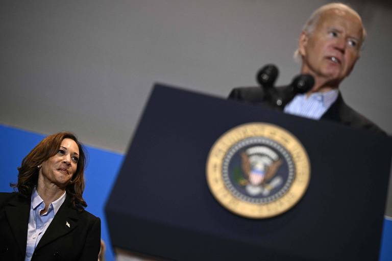 A imagem mostra Joe Biden em um discurso, posicionado atrás de um púlpito com o selo presidencial. À esquerda, Kamala Harris observa atentamente. O fundo é de uma cor azul clara