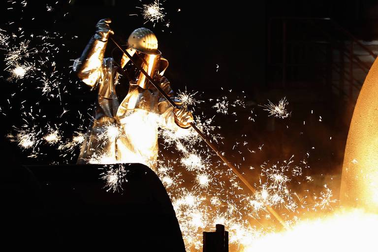 Um trabalhador vestido com um traje de proteção prateado e um capacete, está operando um equipamento na indústria metalúrgica. Ele está usando uma vara longa para manipular metal fundido, enquanto faíscas brilhantes voam ao seu redor, iluminando a cena com um brilho intenso. O fundo é escuro, destacando a atividade e as faíscas.