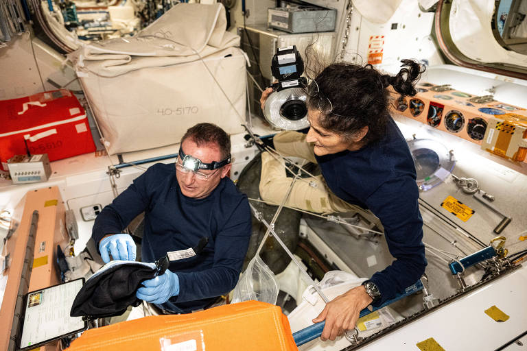 Dois astronautas estão trabalhando dentro da Estação Espacial Internacional. Um astronauta está sentado, usando luvas e um óculos especial, enquanto examina um dispositivo. O outro astronauta está em pé ao lado, observando e segurando um objeto. O ambiente é cheio de equipamentos e ferramentas, com várias caixas e instrumentos ao fundo.