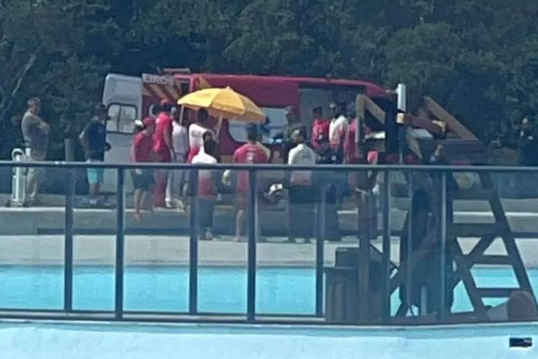 Um grupo de pessoas de vermelho está ao lado de uma ambulância vermelha, próximo a uma piscina azul.