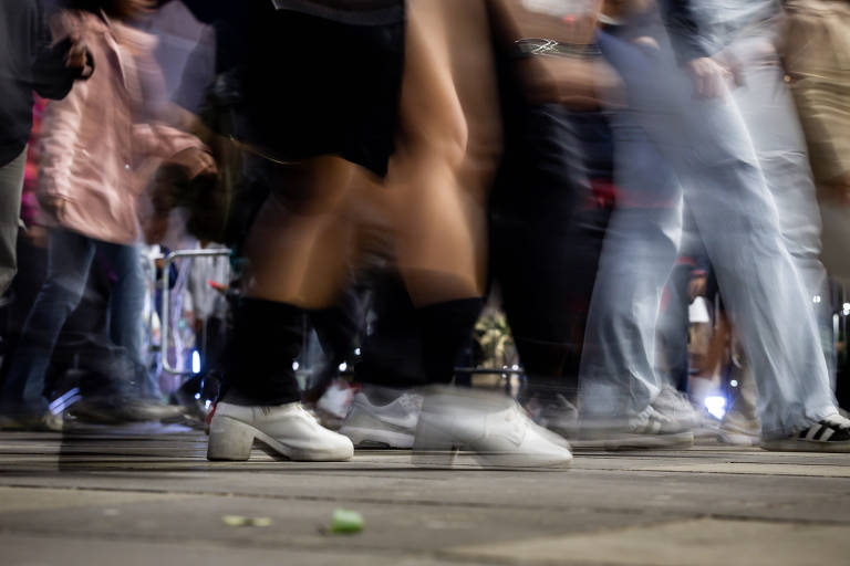 A imagem mostra um grupo de pessoas dançando à noite. As pernas e os pés das pessoas estão em movimento, criando um efeito de desfoque. Algumas pessoas estão vestindo roupas casuais, como camisetas e calças, e há uma variedade de calçados visíveis, incluindo tênis brancos. O ambiente parece ser urbano, com luzes ao fundo.