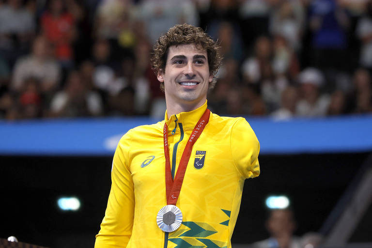 Um atleta masculino, com cabelo cacheado e sorriso, está posando em um pódio. Ele usa uma camisa amarela com detalhes verdes e uma medalha prateada pendurada no pescoço. O fundo mostra uma multidão em um evento esportivo.
