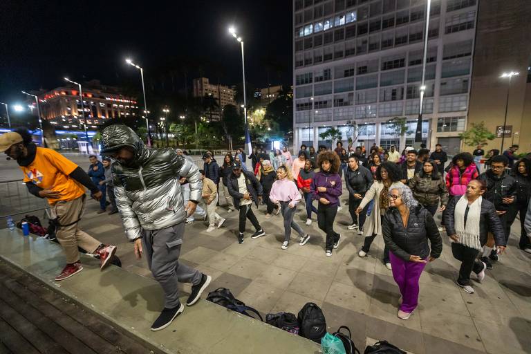 Um grupo de pessoas participa de uma aula de dança ao ar livre durante a noite. Elas estão em um espaço urbano, com prédios iluminados ao fundo. Algumas pessoas estão usando roupas casuais e agasalhos, e a maioria parece estar se divertindo enquanto dança. Há uma variedade de estilos de cabelo e cores de roupas entre os participantes.