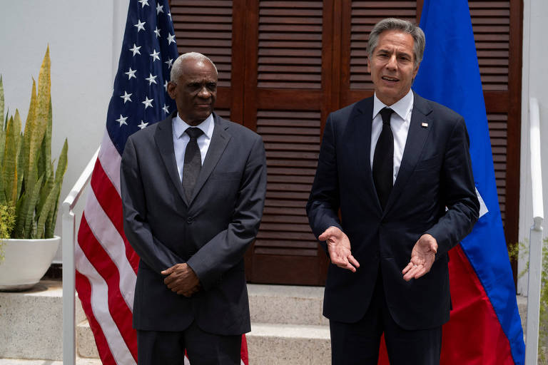 A imagem mostra dois homens em um evento ao ar livre. À esquerda, um homem de pele escura, usando um terno escuro e gravata, está em pé ao lado de um homem de pele clara, que usa um terno escuro e uma gravata preta. Ao fundo, há uma bandeira dos Estados Unidos e uma bandeira do Haiti