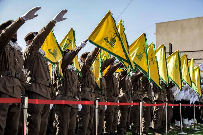 A imagem mostra um grupo de pessoas em uniformes marrons, levantando bandeiras amarelas com um símbolo em verde. Eles estão alinhados em uma formação, com as mãos levantadas em saudação. Ao fundo, há um edifício e o céu está claro.