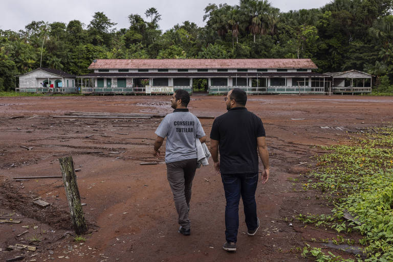 Duas pessoas caminham em direção a um edifício de dois andares, que possui um telhado de duas águas e janelas grandes. O chão é de terra batida e há vegetação ao redor. O céu está nublado e a área parece ser rural ou de floresta.