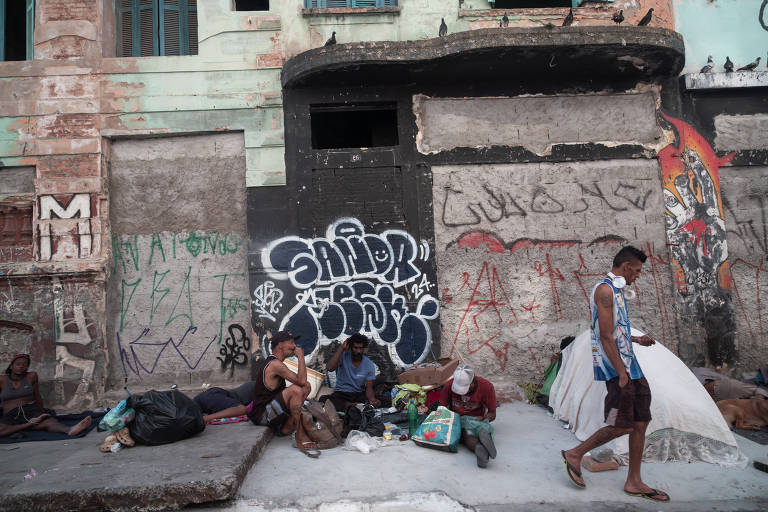 A imagem mostra um grupo de pessoas sentadas em um espaço urbano, próximo a uma parede coberta de grafites. Algumas pessoas estão deitadas no chão, enquanto outras estão em pé ou caminhando. Um homem segura um cachimbo em uma das mãos