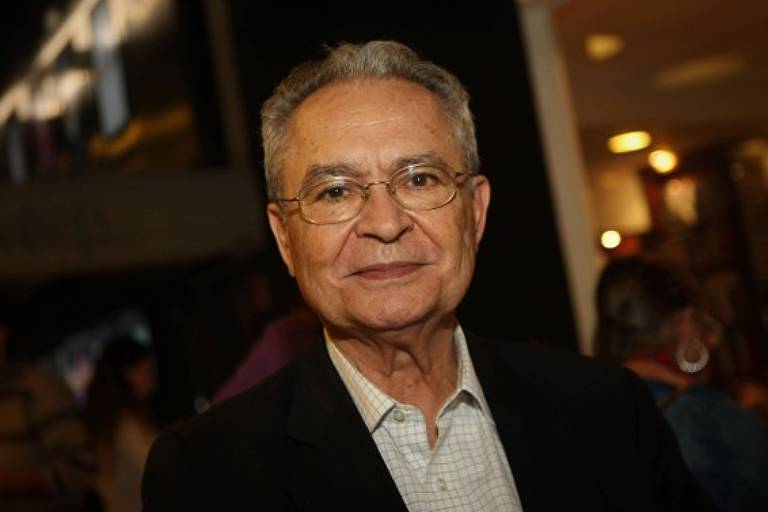 Um homem idoso com cabelo grisalho e óculos, vestindo uma camisa clara e um paletó escuro. Ele está sorrindo levemente e olhando para a câmera. Ao fundo, há pessoas e uma iluminação suave, sugerindo um ambiente social ou de evento.