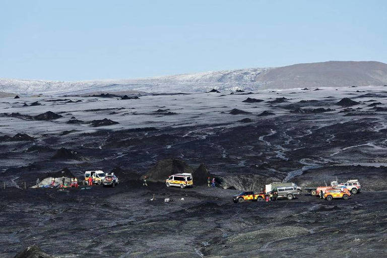 A imagem mostra uma vasta paisagem de gelo escuro. No centro, há um grupo de veículos, incluindo caminhonetes e carros de resgate, estacionados perto da entrada de uma caverna de gelo. Algumas pessoas estão visíveis ao redor dos veículos, e ao fundo, pode-se ver uma extensão de terreno acidentado e montanhas ao longe sob um céu claro.
