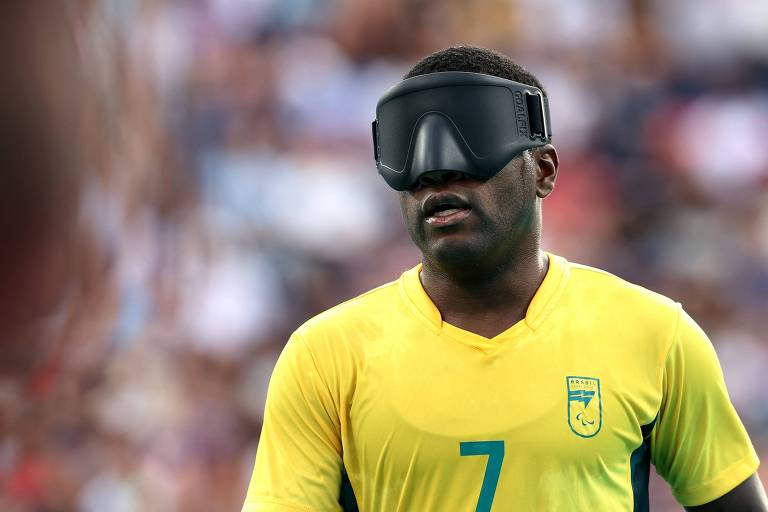 A imagem mostra um atleta vestindo uma camisa amarela com o número 7 estampado, representando o Brasil. O jogador está usando uma venda nos olhos e joga futebol de cegos. Ao fundo, a imagem está um pouco desfocada, com um público assistindo ao evento