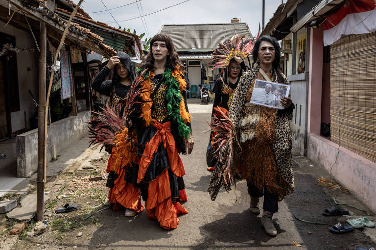 A imagem mostra um grupo de pessoas vestidas com trajes coloridos e elaborados, caminhando por uma rua estreita. Os trajes incluem penas, tecidos vibrantes em laranja e verde, e acessórios que parecem ser feitos de materiais naturais. Ao fundo, há casas simples e algumas pessoas observando. O céu está nublado, sugerindo um dia sem sol