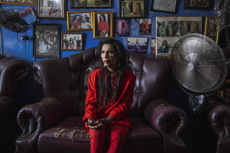 A imagem mostra uma mulher sentada em um sofá de couro marrom, vestindo um conjunto vermelho. Ela está em um ambiente com paredes azuis decoradas com várias fotos emolduradas. Ao lado, há um ventilador grande. A iluminação é suave, criando uma atmosfera íntima