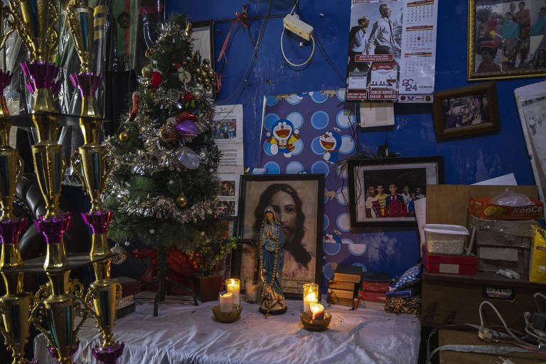 A imagem mostra um altar decorado com uma pequena árvore de Natal à esquerda, adornada com enfeites coloridos. No centro, há uma imagem de uma figura religiosa emoldurada, cercada por velas acesas. O fundo é azul e há várias fotos e documentos pendurados na parede, além de caixas e livros empilhados na parte inferior direita