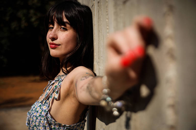 A imagem mostra uma mulher com cabelo escuro e liso, usando um vestido floral. Ela está posando de lado, com um leve sorriso e olhando para a câmera. Sua mão está estendida em direção à lente, destacando um esmalte de unha vermelho. O fundo é desfocado, com árvores e um chão de terra visíveis.